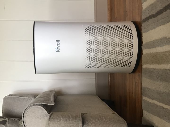 A white tower air purifier standing on a wooden floor beside a sofa
