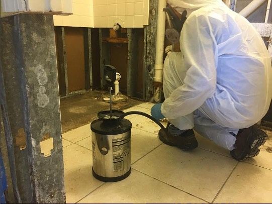 A technician in a protective suit working at an open floor duct with a sprayer